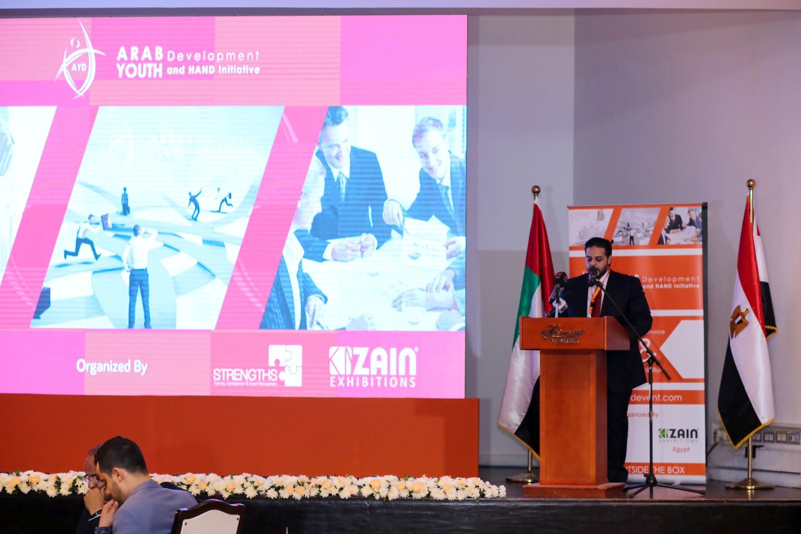 Speaker at the AYD 2016 podium; stage screen shows the Arab Youth Development branding, organised by Strengths and Zain Exhibitions